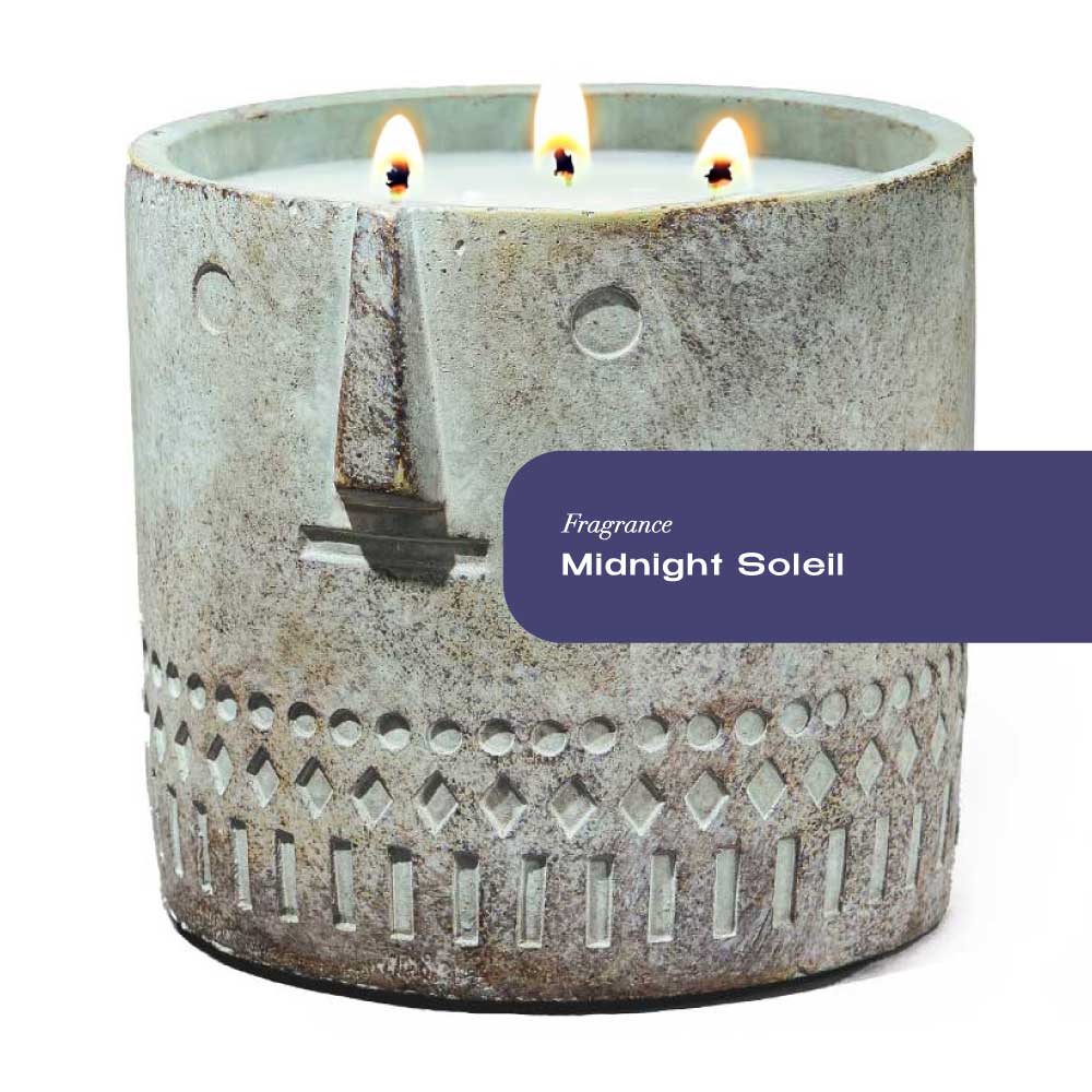 Hand-painted cement vessel candle with a serene stone face design, glowing warmly in dim light.