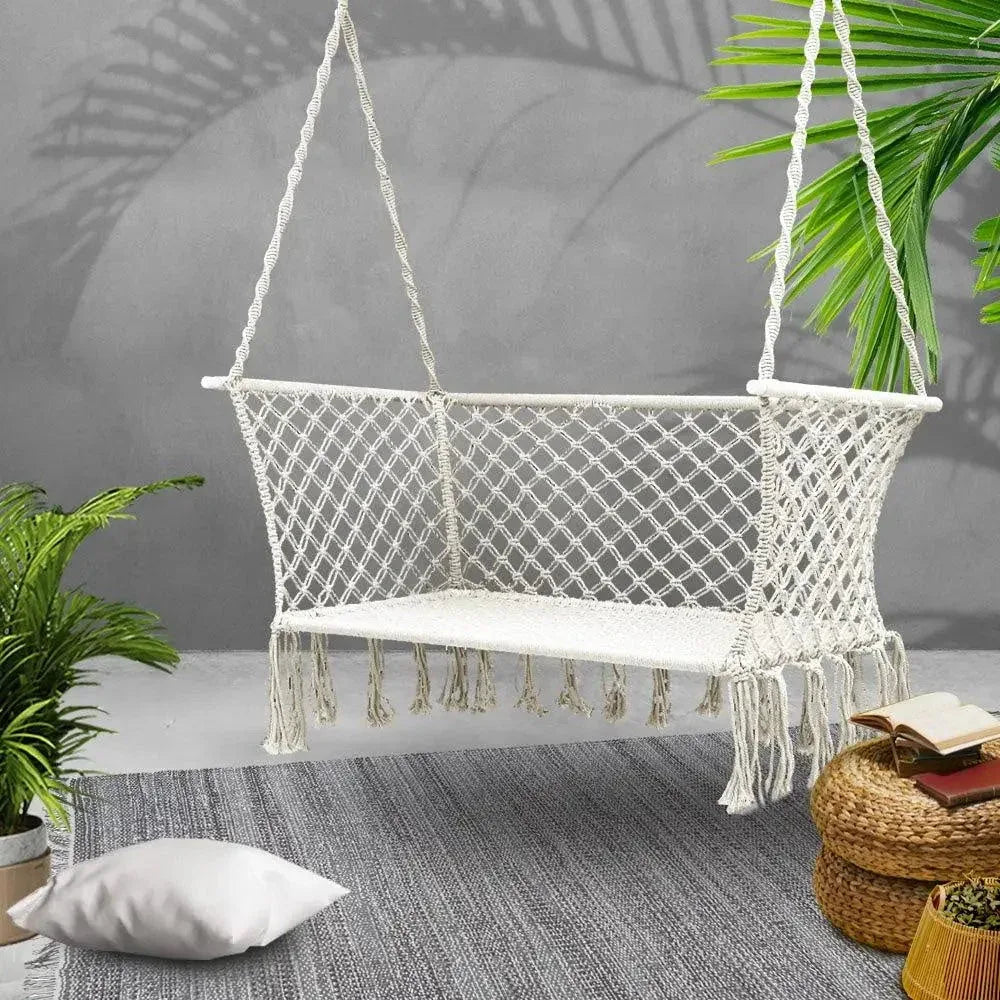 Two-person hammock chair with tassels hanging outdoors next to a tree.