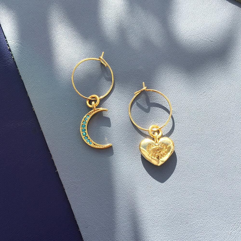 Close-up of turquoise crystal moon and chunky gold heart mini hoop earrings on white background.