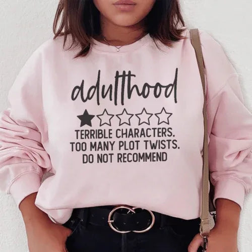 Light pink sweatshirt featuring the phrase 'Adulthood Terrible Characters Too Many Plot Twists' in bright lettering