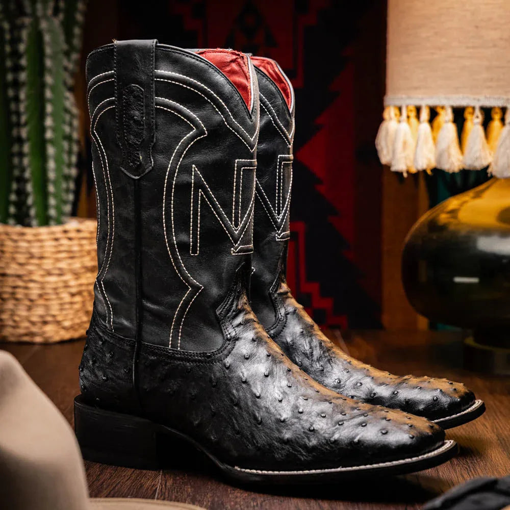 Man wearing black ostrich skin cowboy boots standing outdoors on rocky terrain.