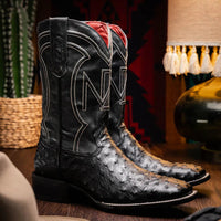 Man wearing black ostrich skin cowboy boots standing outdoors on rocky terrain.