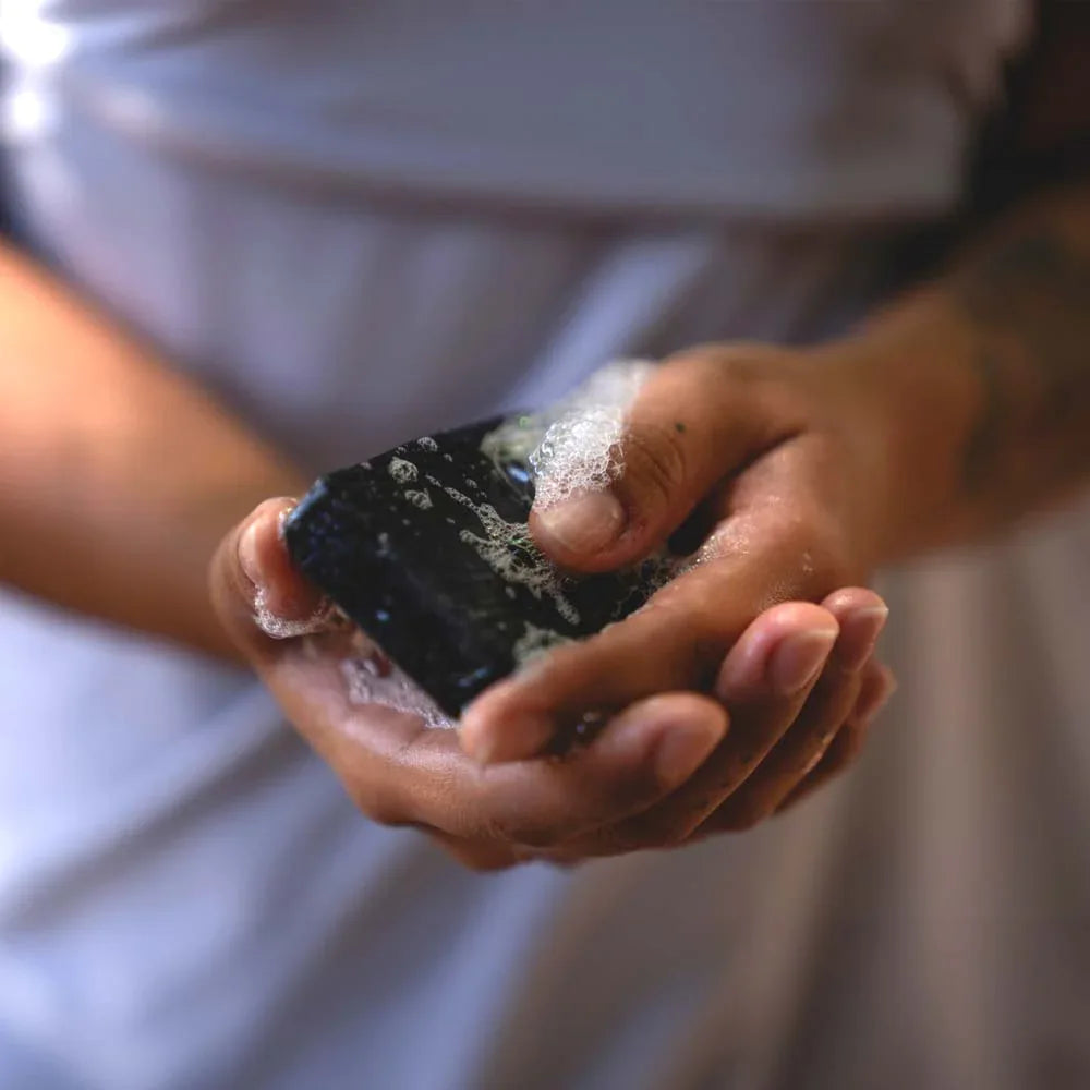 Hand holding a smooth, dark charcoal bar of organic tea tree activated charcoal soap.