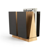Standing magazine holder WINGS in oak wood and black metal with magazines arranged inside on a wooden floor.