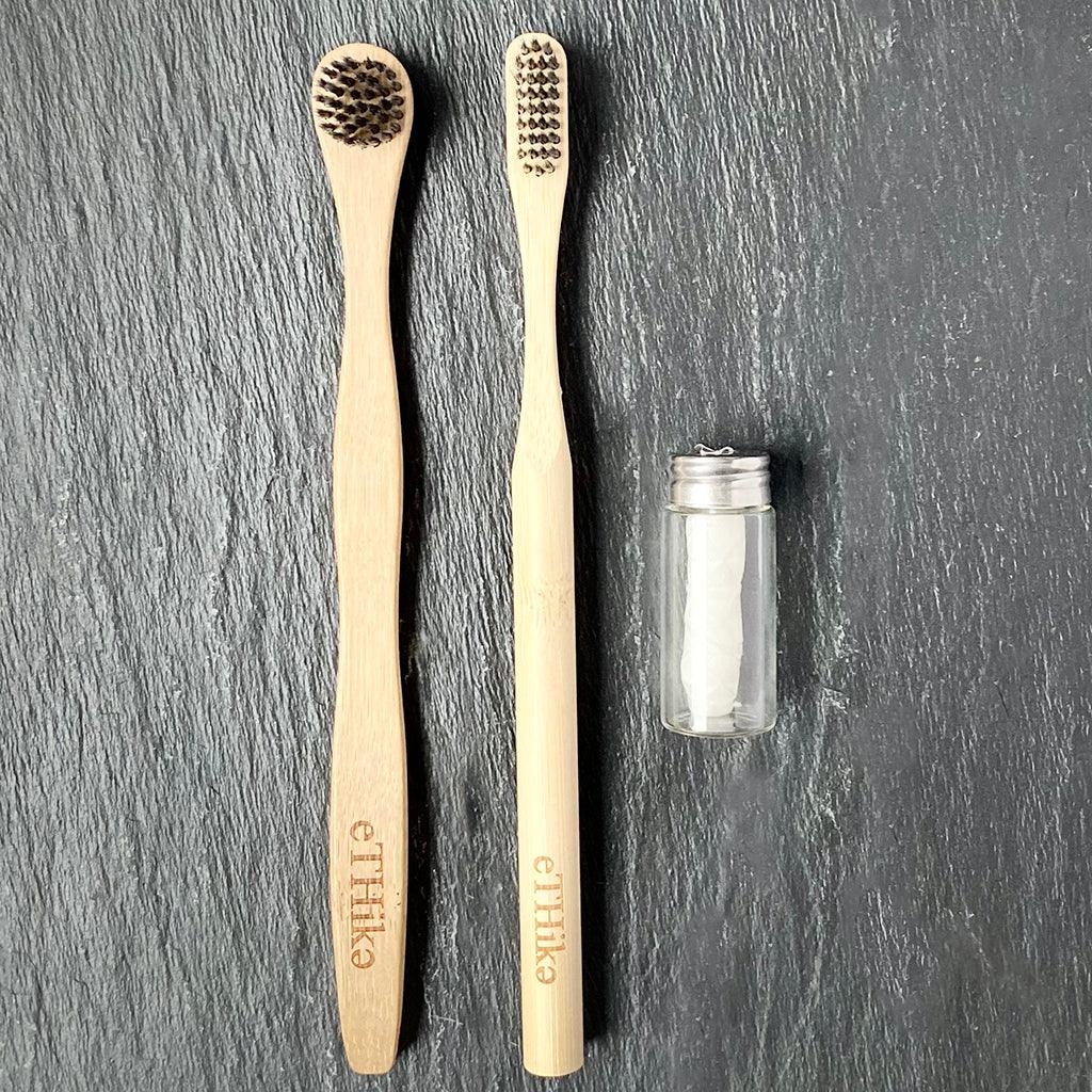 Close-up of bamboo toothbrush with charcoal bristles alongside tongue brush and glass jar dental floss.