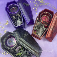 Resin coffin-shaped tealight candle holder with embedded rose quartz, dried flowers, and a skeleton hand design.