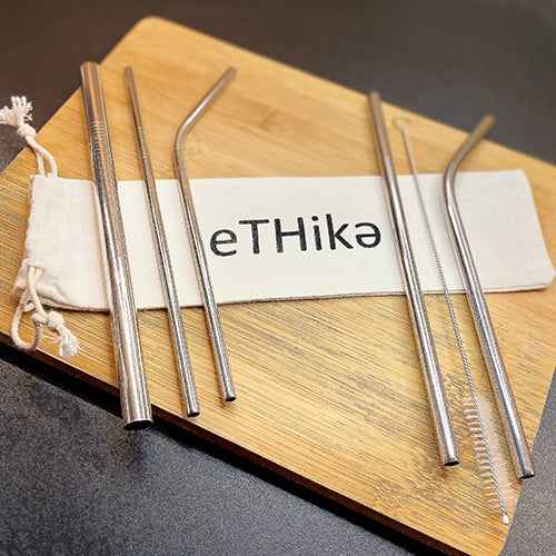 Eleven stainless steel straws displayed alongside a white cotton pouch and cleaning brush on a wooden surface.