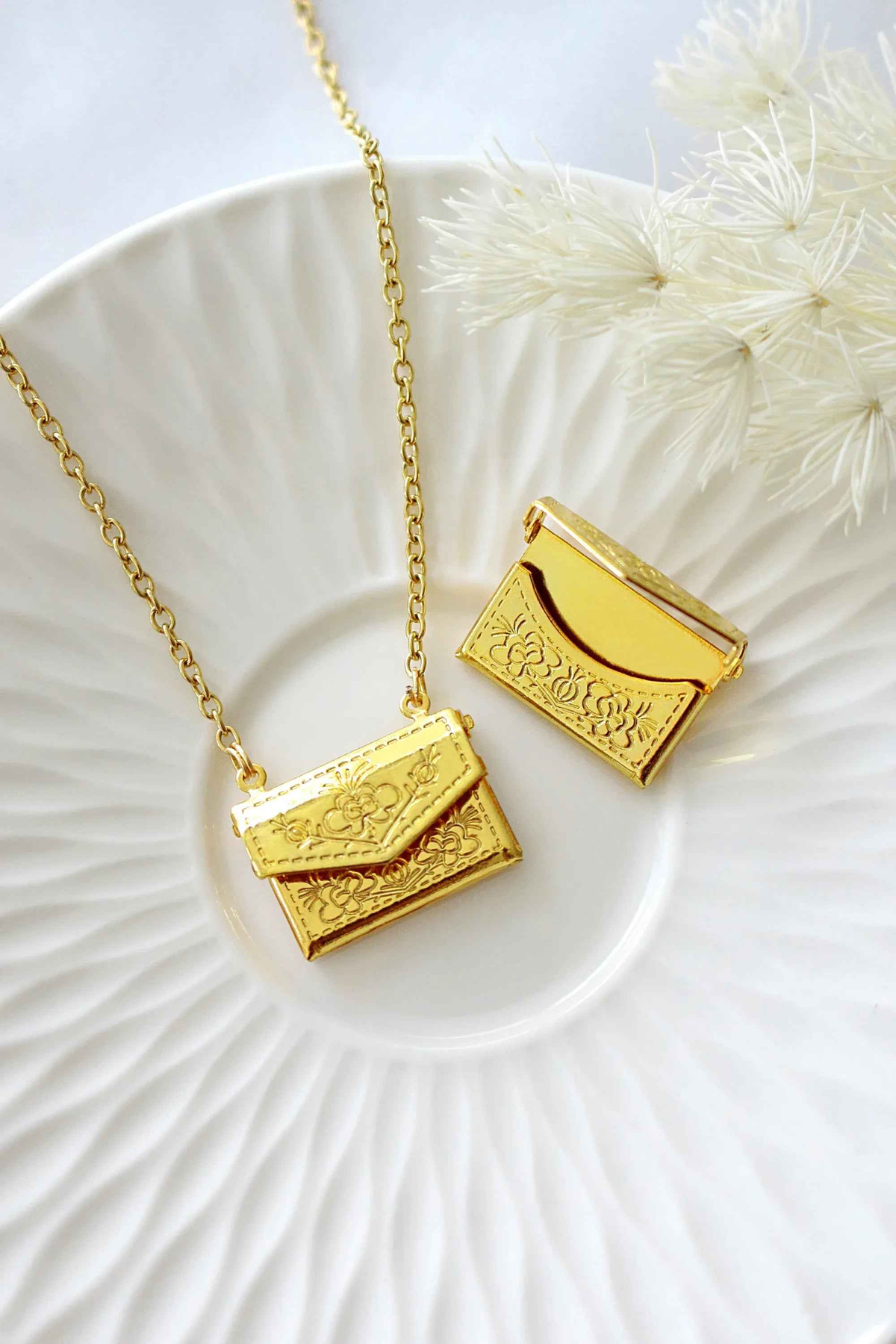 Gold-plated envelope locket necklace with delicate daisy engraving, displayed on a soft neutral background.
