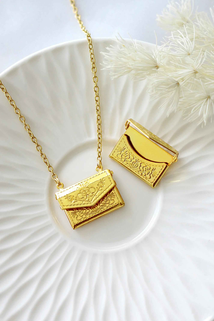 Gold-plated envelope locket necklace with delicate daisy engraving, displayed on a soft neutral background.