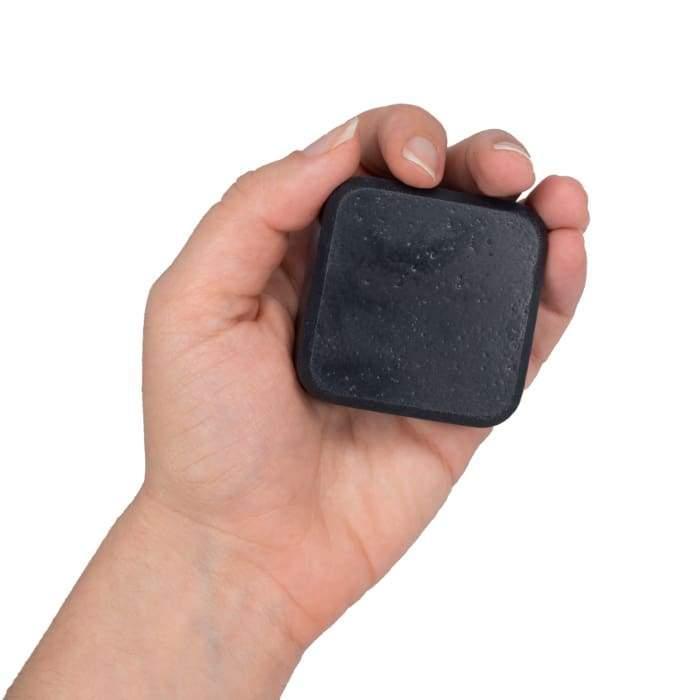 Charcoal facial soap bar alongside a finger wallet and electronic device on a neutral surface.