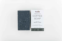 Close-up of charcoal coffee soap bar on a wooden surface with scattered coffee beans and a cloth background.