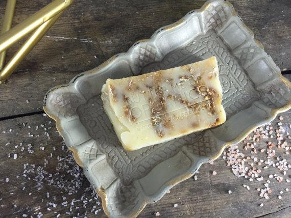 Oat and honey soap bar with textured surface on wooden soap dish with honey and oats nearby.
