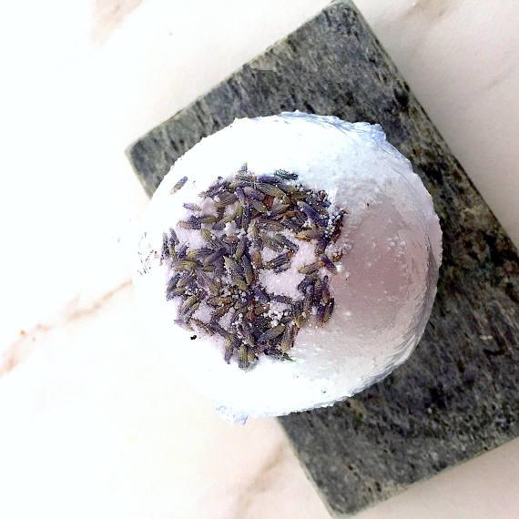 Hand holding a round lavender bath bomb with visible natural ingredients embedded.