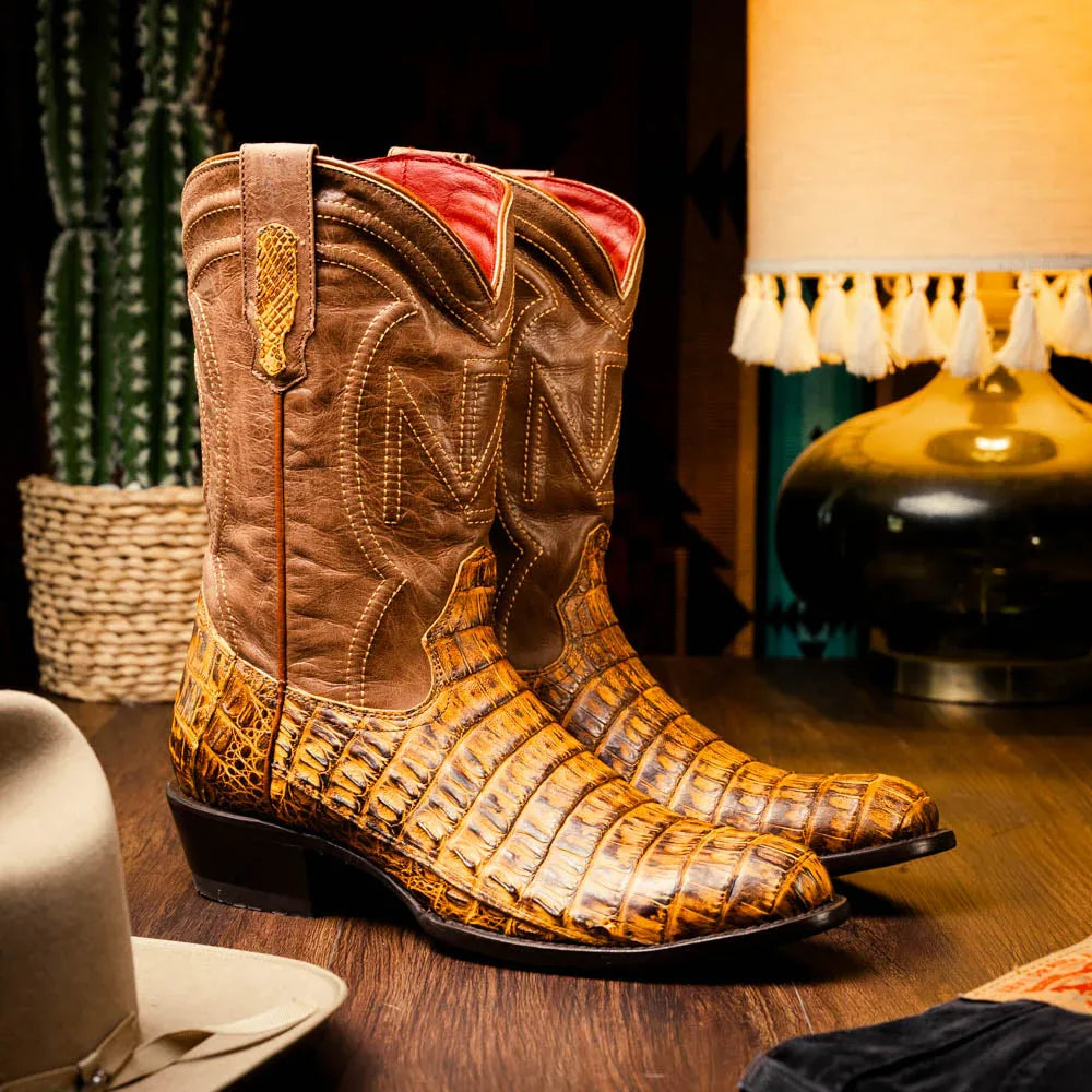 Man wearing mustard brown genuine caiman belly skin cowboy boots standing on wooden floor.